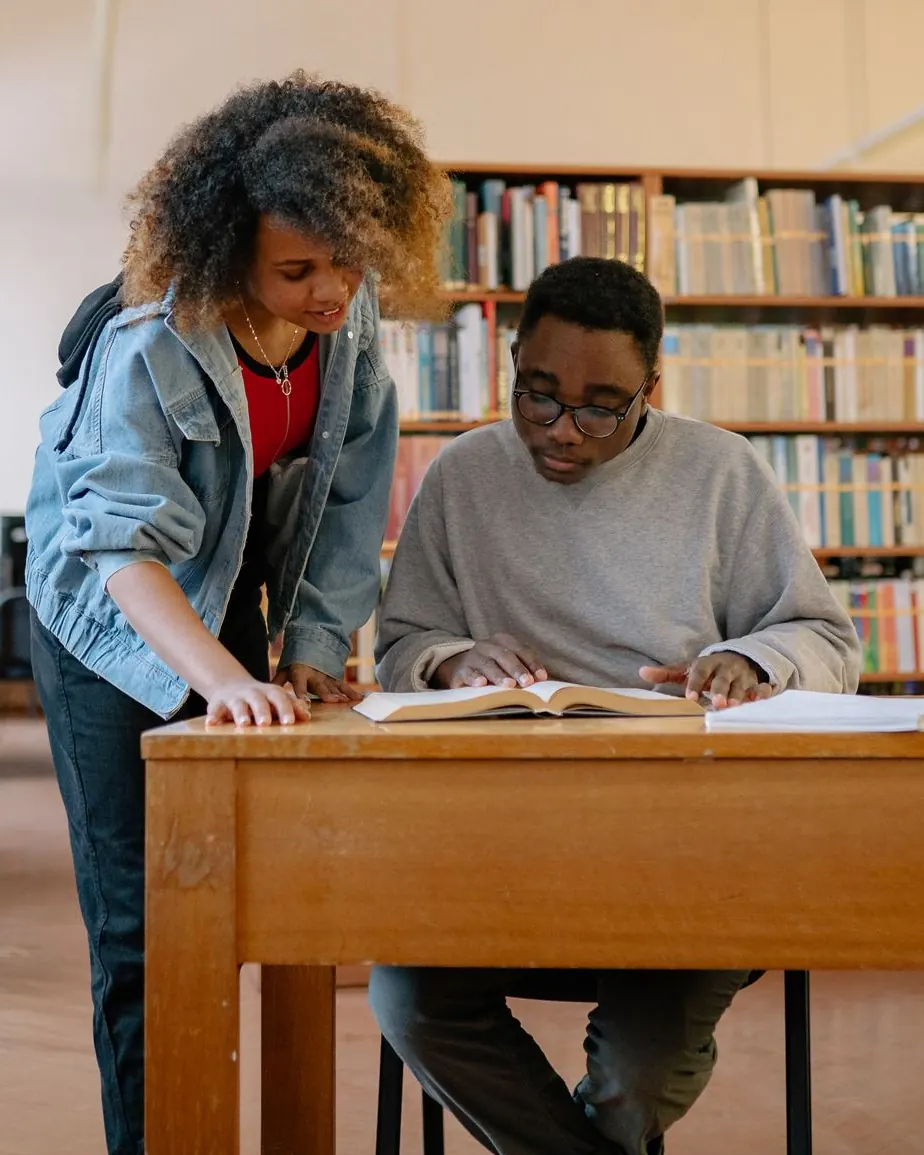 A student mentor leaning over a desk to guide another student through their coursework in a library